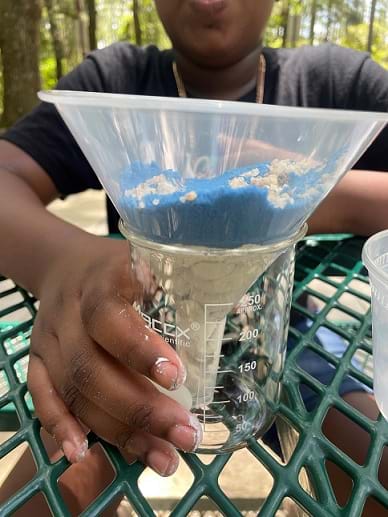 A child’s hand holds a glass beaker with a funnel placed inside. The funnel is clear and white clay, blue sand, and small pebbles are layered within.