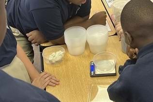 Four students gather around a desk with a digital scale, stacks of empty containers, and a container with air dry clay in it. There is an empty container placed on the scale as the teacher opens the container of a material and students are watching.