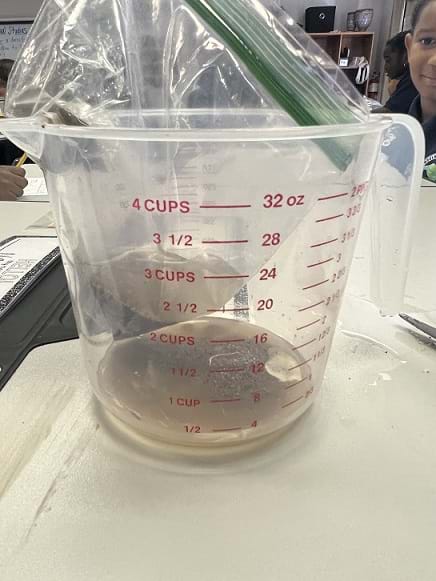A small corner of the plastic bag was cut, and water was poured through the bag. Water drains through the soil and gets deposited in the measuring cup. 