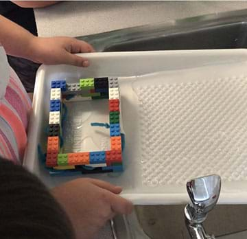 The photo shows a paint tray holding a house made of plastic bricks. Water is flowing into the tray. The plastic house has pipe cleaners and tape protruding from two windows.  