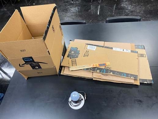 Activity materials are on a table: a cardboard box, a small pile of flat pieces of cardboard, and a lightbulb in a lightbulb base. 