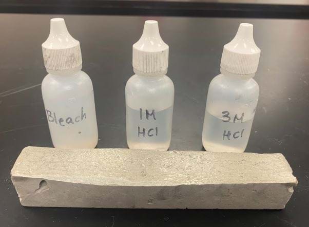 The image shows the acid vs. base drop test station. Concentrated bleach and different concentrations of Hydrochloric acid are placed in dropper bottles. One drop of each is placed on different parts of the concrete sample one at a time. This is used to test the reactivity of the concrete to strong acids and strong bases.