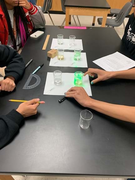 Students perform the experiment. One student shines a laser through a glass of water, and another student observes the experiment. 