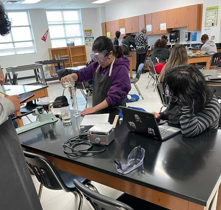 A student wearing lab goggles and an apron fills a graduated cylinder.