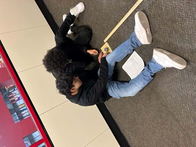 Two students prepare to measure force. The students are placing a weight on top of a block of wood. A meter stick is next to the block of wood. 