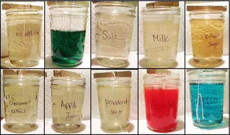 A composite image shows side-view photographs of 10 Mason jars filled with liquids into which white cotton strings hang from clothespins resting across the jar mouths. The "no additive" (control) jar is a clear liquid. The nine jars with additives contain food coloring (green liquid), salt (clear), milk (slightly cloudy white), brown sugar (light brown), cinnamon extract (clear), apple juice (clear), powdered sugar (clear), Kool-Aid (cloudy red), JELL-O (transparent blue).