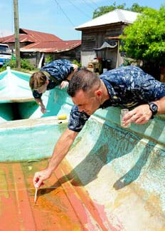 Photograph shows two men leaning into a small boat on dry land, gathering samples from the shallow amount of orange-colored water in the bottom of the boat. They work as part of a humanitarian and civil mission looking for contaminated water in Guatemala.