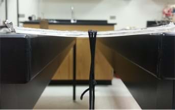 A photograph shows two tables placed a few inches apart, on which the opposite ends of a long, clear mold of an epoxy and amine mixture rest. A weighted string hangs from the center of the mold, which is bowed a little, showing the flexibility that exists under strain.