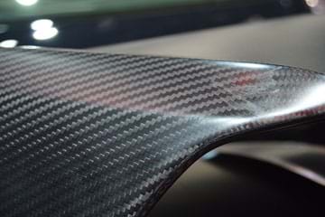 A close-up photo shows a car spoiler made of curved carbon-fiber-reinforced polymer. It looks like a curvy black plastic material with a smooth but visible woven gray/black pattern.