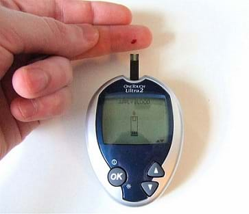 A photograph shows a hand with a blood spot (sample) on the pad of the end of one finger positioned near a computer mouse-shaped plastic device with buttons, a display screen and a probe sticking out towards the finger. 