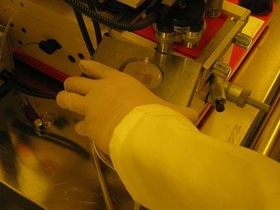 A photograph shows a technician's gloved hand placing a prepared wafer where a circuit is going to be printed in a piece of equipment (a Mask Aligner-UV).