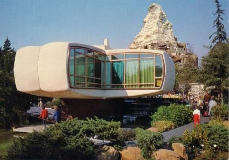 A photograph shows a disc-shaped white house sitting atop a large post. The house is made of plastic, and a portion of the disk-shape is cut-away (like a removed pie piece) with glass windows so the inside contents may be viewed. The house is located amidst greenery with a stone mountain peak behind it.