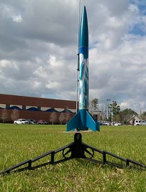 A photograph shows a model blue rocket ready to launch. The rocket sits on a grassy field next to a building.