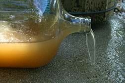 A photograph shows brownish, cloudy dirty water spilling from a clear glass gallon jug lying on its side. A photograph shows brownish, cloudy dirty water spilling from a clear glass gallon jug lying on its side.