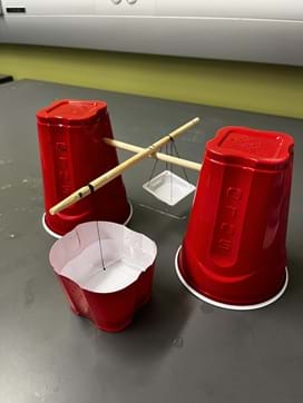 A photo showing the balance for the Method 1 activity. It is a balance beam made from plastic cups, wooden dowels, thread, needle, and weigh basket.  A photo showing the balance for the Method 1 activity. It is a balance beam made from plastic cups, wooden dowels, thread, needle, and weigh basket.