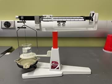 A photo showing the balance for the Method 2 activity. It is a modified triple-beam balance with a suspended button and a beaker. A photo showing the balance for the Method 2 activity. It is a modified triple-beam balance with a suspended button and a beaker.