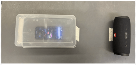 A smartphone displaying a decibel app inside a clear plastic container, next to a black JBL Bluetooth speaker, set up for a sound experiment.