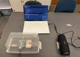 A photo of a complete sound insulation experiment setup. A laptop displays code, controlling a sound detection circuit (Arduino and breadboard) enclosed in a plastic container, while a Bluetooth speaker provides the sound source.