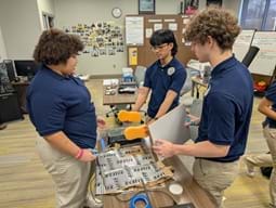 A photo of three students working at a table, examining materials for the activity, with one holding a white board and another student manipulating items on the table.