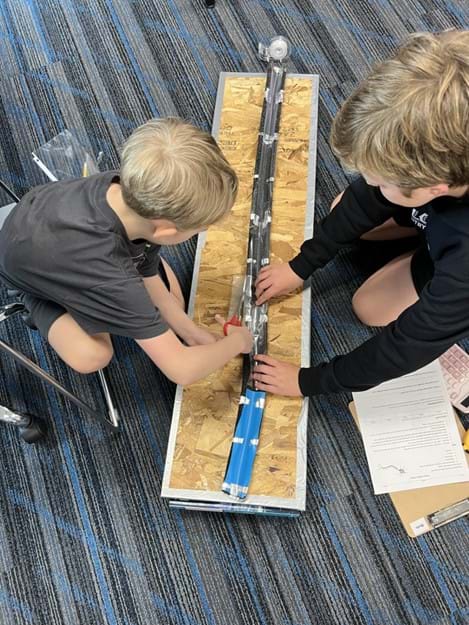 Two students adding plastic material to a foam tube. One student is cutting plastic using the foam tube as a size guide, while the other is holding the plastic to keep it stable for cutting.