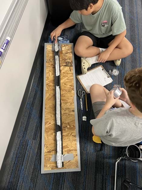 A photo showing two students seated next to a foam tube covered in aluminum material with missing sections of aluminum. One student is about to release a marble into tube, while the other is about to unroll tape to make repairs.