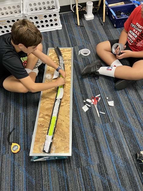 A photo showing two students seated next to a foam tube covered in aluminum and rubbery material. One student is attaching new pieces to the foam to replace damaged pieces, while the other student is about to unroll tape.