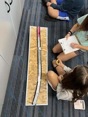 A photo showing two students seated next to a foam tube covered in aluminum and rubbery material. One student is writing on a data sheet, while the other is holding a stopwatch.