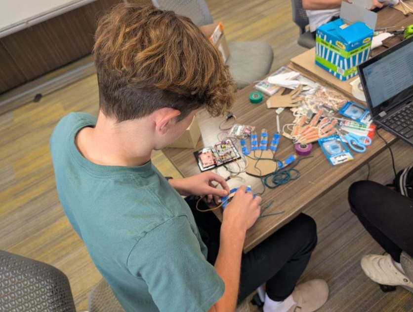 A photo showing a student sitting at a table connecting the wires between his cardboard hand design to the Arduino circuit board.