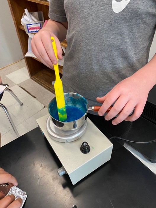 A photo showing a student using a plastic stir stick to mix up the gelatin-based bioplastic recipe. The mixture is blue and is being melted in a pot on a hot plate.