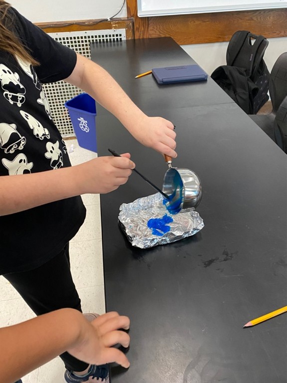 A photo showing a student pouring the blue mixture out of the pot onto a piece of aluminum foil to be flattened and cooled.