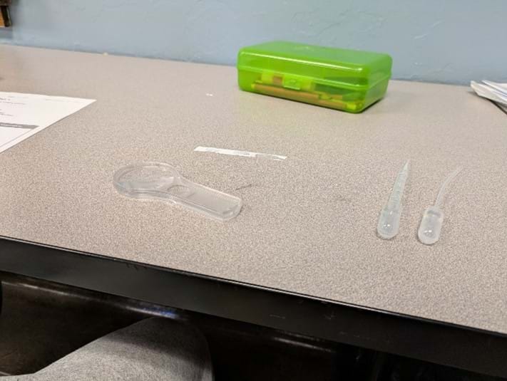 A photo showing a strip of Teflon tape with some water drops along with two pipettes and a magnifying glass on a worktable.