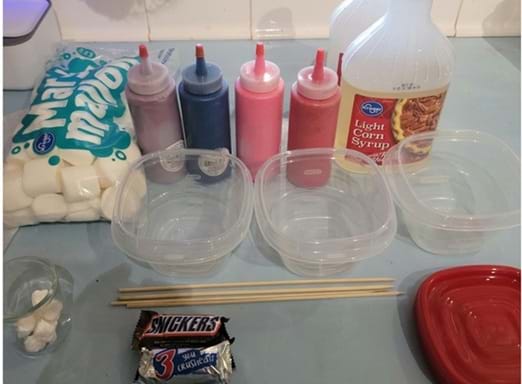A photo showing materials for a candy-dipping activity. On the left are a bag of large marshmallows and four squeeze bottles containing different colored liquids. Behind the containers is a bottle of light corn syrup. In front are three empty clear plastic containers, two wooden skewers, a small pile of cotton or paper pieces, and a fun-size Snickers candy bar. 