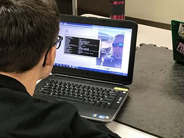 A young man in glasses working on a laptop computer, extracts metadata from a picture of an older man in sunglasses.