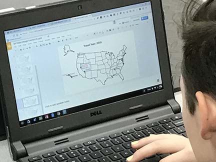 A young man at a laptop computer, is working on a PowerPoint presentation, inputs visual representation of positional data on an image of the United States.