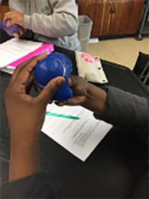 One student in foreground pushing a balloon filled with hydrogels through a shower ring, simulating the blebbing action as the nucleus of a cell migrates through a stiff ECM.