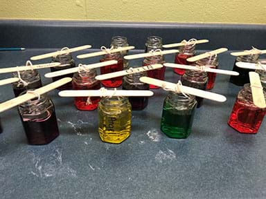 Multiple small glass jars filled with colored water with a large popsicle stick laying across the top of each jar. 