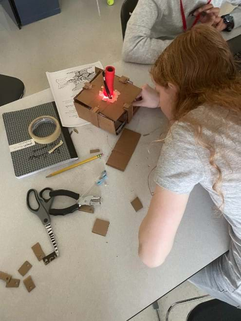 A student is observing how her product, a laser maze, works by shining a flashlight into the maze and seeing where the light hits the mirrors.