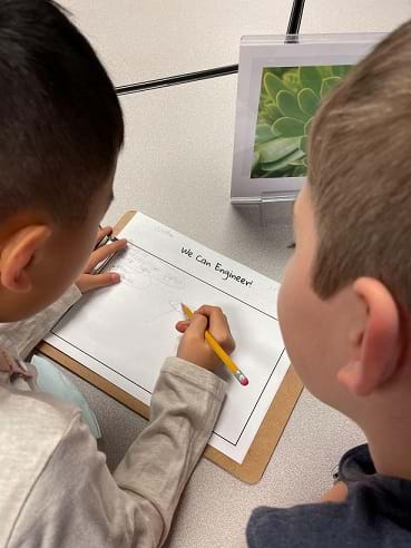 Two students stand over a worksheet on a table. One student is holding a pencil above the paper. 