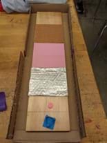 Student prototype with a board modified with four surfaces (wood, bubble wrap, foam, sandpaper, wood). A launcher is placed at the front of the board that the player can use to launch the puck. The launcher uses a rubber band slingshot mechanism.