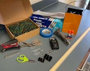 A photo of a box of foil sheets, stapler, staples, a pair of scissors, blue painter’s tape, a box of marbles, orange bucket, sheet metal vise grips, two glue sticks, a meter stick, two large binder clips, a hole punch, rubber bands, and a hanging scale. 