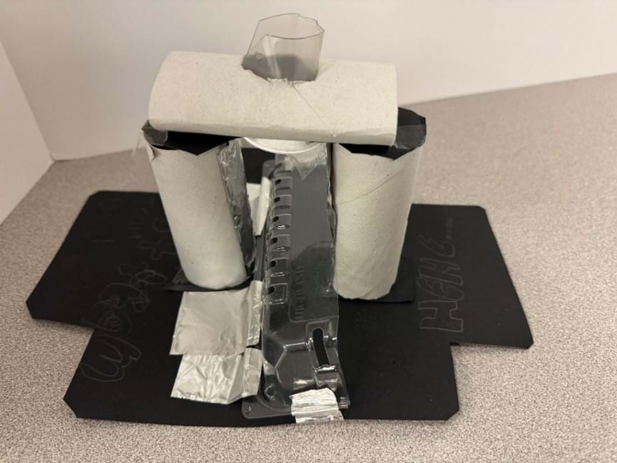 A photo of a DIY microscope model constructed using various household and craft materials. The base of the microscope is made from black cardstock, with visible handwritten text and doodles in white crayon. Two cardboard tubes, from toilet paper rolls, stand vertically as structural supports on each side of the central microscope body. The main body is a repurposed clear plastic container, from food packaging, with silver duct tape used to hold it in place and connect parts. A top horizontal cardboard toilet paper roll bridges the two vertical tubes and supports a transparent plastic piece, which houses the lens or eyepiece. The overall model resembles the general shape of a compound microscope.