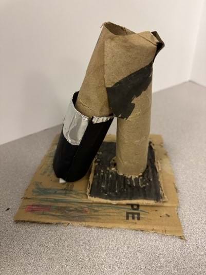 A photo of a DIY microscope model constructed primarily from cardboard materials is displayed on a flat surface. The base consists of a square piece of corrugated cardboard that has been drawn on with various colors, including red, green, and black. Two cardboard tubes are attached vertically to the base—one taller and slightly bent at the top, possibly representing the microscope's eyepiece, and another shorter tube wrapped in black paper and silver duct tape, likely symbolizing the objective lens. Black paint or marker has been used to add details on the tubes and base, giving it a more realistic microscope appearance.