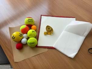 A photo showing several types of balls (tennis balls, ping pong balls, a stress ball, and a foam ball) and a rolled-up tape measure. The items are sitting on a few different surfaces that are fanned out: a sheet of sandpaper, a small dry erase board, and sheet of foam.
