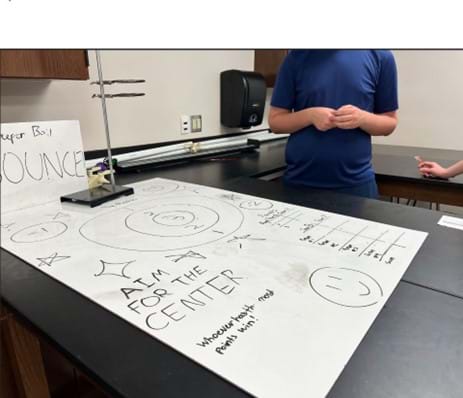 A photo showing a student-created game with a whiteboard with a target board drawn on it and science lab equipment surrounding it.