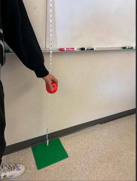 A photo showing a red wiffle ball held over a green folder, about to be dropped. A measuring tape hangs on the wall behind the ball.