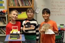 A photo of three students standing, each holding a different model house made from different materials. 