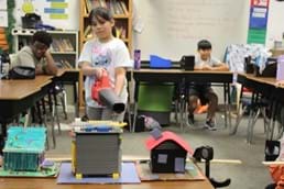A photo showing a student pointing a leaf blower at three model houses while two students sit behind their desks watching. 