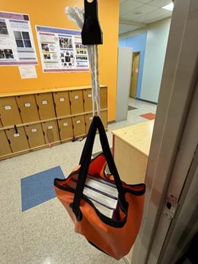 A photo of a classroom scene showing a hanging test of paper-strap prototypes. A black spring-scale-style clip at the top suspends three twisted white paper strands that are tied to the black fabric handles of an orange tote bag. The bag is filled with several thick hardcover books and hangs in a doorway above linoleum floor tiles. In the background are wooden cubbies, colorful wall posters, and a light-blue hallway. A photo of a classroom scene showing a hanging test of paper-strap prototypes. A black spring-scale-style clip at the top suspends three twisted white paper strands that are tied to the black fabric handles of an orange tote bag. The bag is filled with several thick hardcover books and hangs in a doorway above linoleum floor tiles. In the background are wooden cubbies, colorful wall posters, and a light-blue hallway.