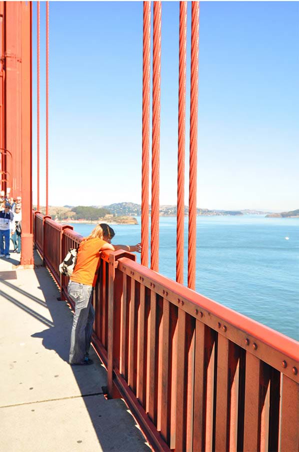 A photograph of a close-up view of the steel suspender ropes of the Golden Gate Bridge. These vertical ropes, spaced 50 feet apart, connect the bridge's main cables to the roadway deck. The image includes a person leaning on a railing of the bridge and looking out toward the bay. A photograph of a close-up view of the steel suspender ropes of the Golden Gate Bridge. These vertical ropes, spaced 50 feet apart, connect the bridge's main cables to the roadway deck. The image includes a person leaning on a railing of the bridge and looking out toward the bay.