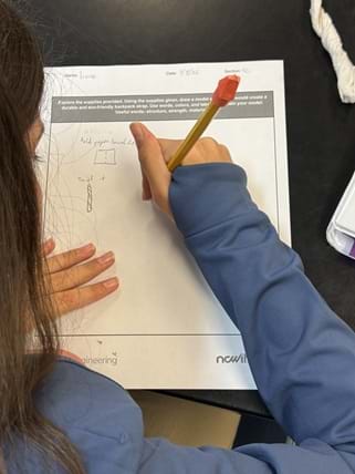 A photo taken over the shoulder of a person seated at a black tabletop, drawing on a printed worksheet with their right hand. The worksheet has printed text and small hand-drawn sketches. A crumpled paper towel is at the top right, and a white phone or case is partly visible at the right edge. A photo taken over the shoulder of a person seated at a black tabletop, drawing on a printed worksheet with their right hand. The worksheet has printed text and small hand-drawn sketches. A crumpled paper towel is at the top right, and a white phone or case is partly visible at the right edge.